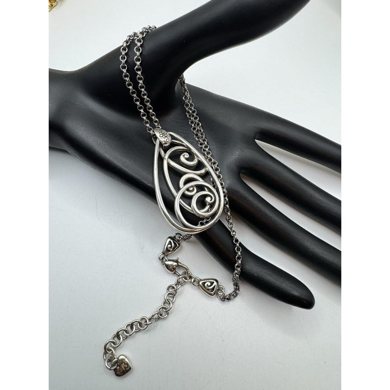 May include: Silver-toned pendant necklace with an open teardrop design. The pendant features an intricate, swirling pattern within the frame. The necklace has a delicate chain with heart-shaped accents near the clasp and a heart-shaped charm at the end.