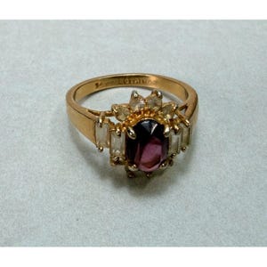 Op de afbeelding: Een gouden ring met een centrale ovaal geslepen dieppaarse edelsteen, omringd door kleinere heldere baguette- en rond geslepen stenen. De binnenkant van de ring is gestempeld met de tekst 14K GESTAIWAN, wat de zuiverheid en herkomst aangeeft.