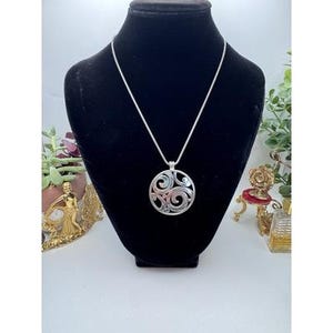 以下が含まれることがあります： 円形デザインのシルバーペンダントネックレス。ペンダントは渦巻き模様のオープンワークパターンが特徴です。ネックレスは黒いマネキンに飾られており、ジュエリーのデザインと詳細が際立っています。チェーンは細いシルバーカラーのスネークチェーンです。