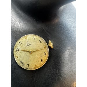 May include: A vintage gold-toned Omega watch face with black numerals and hands. The watch face has a textured surface and a small crown on the side. The Omega logo is visible at the top.