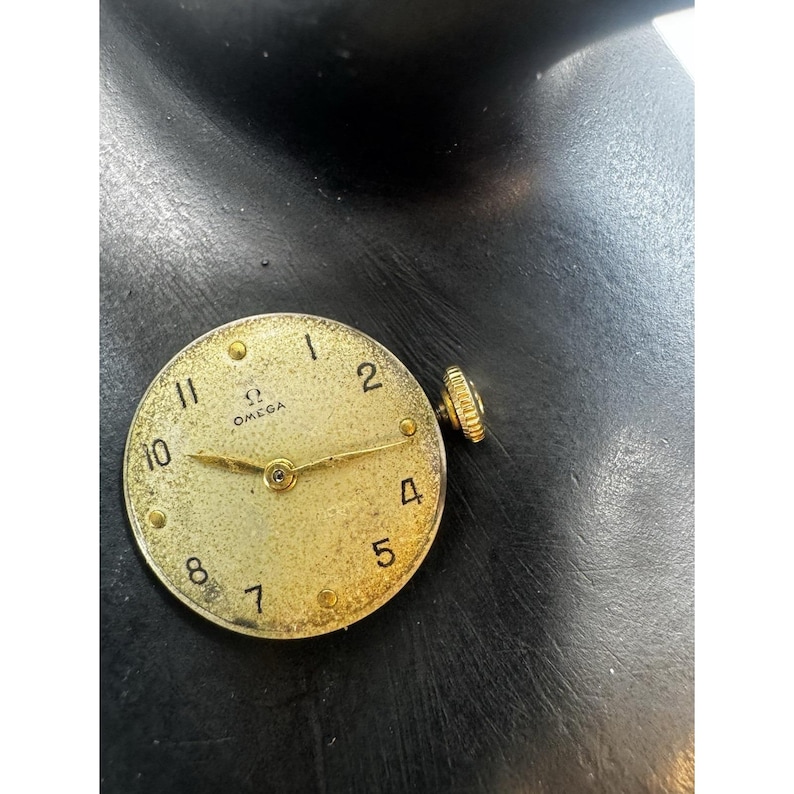 May include: A vintage gold-toned Omega watch face with black numerals and hands. The watch face has a textured surface and a small crown on the side. The Omega logo is visible at the top.
