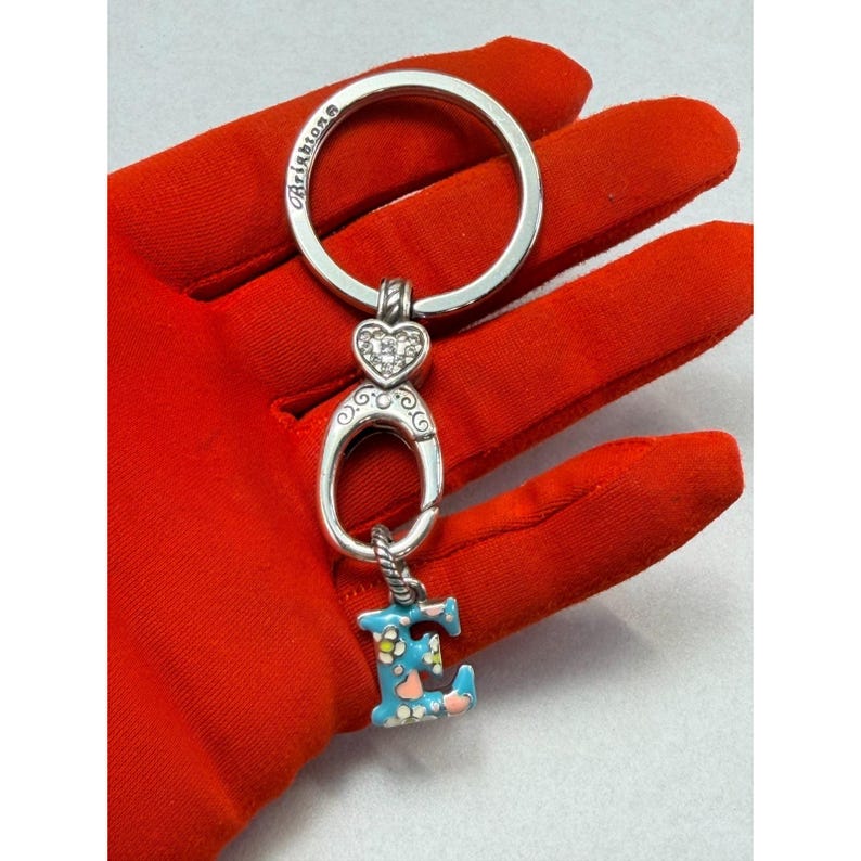 Peut inclure: Porte-cl&eacute;s argent&eacute; avec un grand anneau, un charm en forme de c&oelig;ur orn&eacute; de pierres transparentes et une lettre "E" en &eacute;mail bleu orn&eacute;e de fleurs. Le porte-cl&eacute;s est pos&eacute; sur un fond rouge.