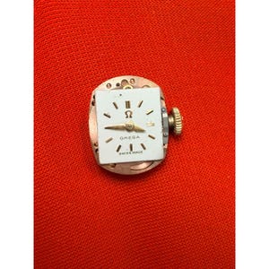 May include: A vintage Omega watch movement. The white watch face has black markers, gold hands, and the Omega logo. The words "OMEGA" and "SWISS MADE" are printed on the face. It has a gold crown and is set on a copper-coloured base.