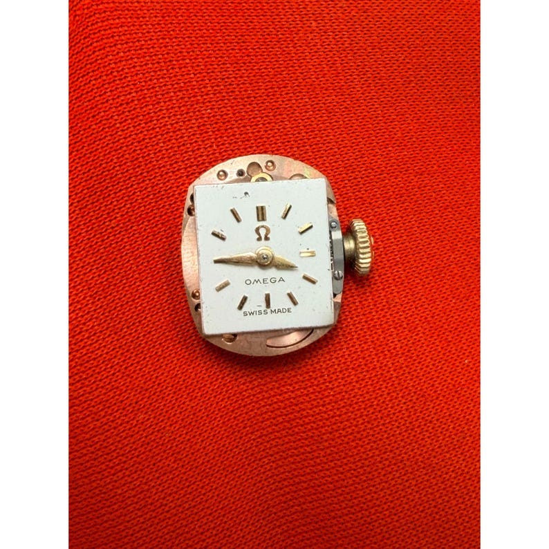 May include: A vintage Omega watch movement. The white watch face has black markers, gold hands, and the Omega logo. The words "OMEGA" and "SWISS MADE" are printed on the face. It has a gold crown and is set on a copper-coloured base.