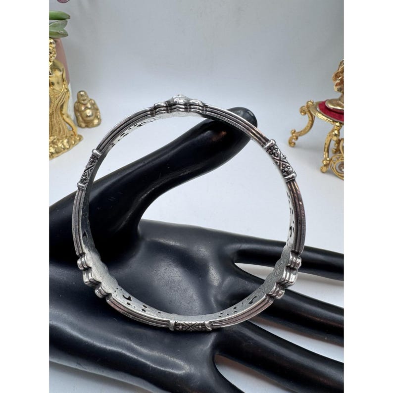 May include: A silver-toned bangle bracelet with intricate floral and geometric patterns. The bracelet is round and appears to be made of metal. The design includes detailed carvings and a textured surface, showcasing a vintage or antique style.