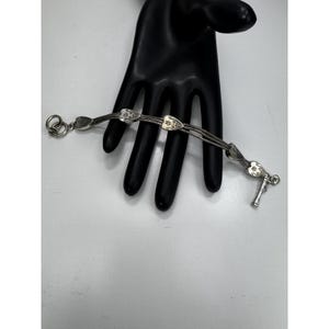 May include: A silver bracelet with a delicate design. The bracelet features three strands of linked chain, adorned with heart-shaped charms and floral details. The clasp is a T-bar style. The bracelet is displayed on a black hand-shaped display.