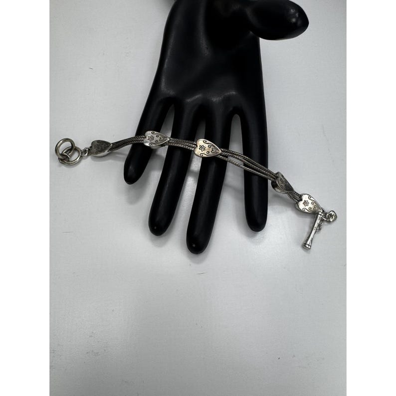 May include: A silver bracelet with a delicate design. The bracelet features three strands of linked chain, adorned with heart-shaped charms and floral details. The clasp is a T-bar style. The bracelet is displayed on a black hand-shaped display.