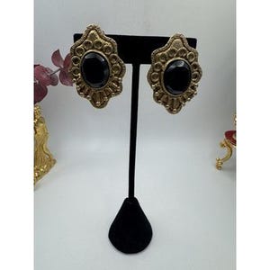 May include: A pair of vintage gold-tone clip-on earrings with a black oval center stone. The earrings have an ornate, filigree design around the stone. They are displayed on a black earring stand.