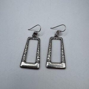 May include: A pair of silver-toned rectangular earrings. The earrings have a geometric design along the edges and a solid bar at the bottom. The hooks are curved and simple.
