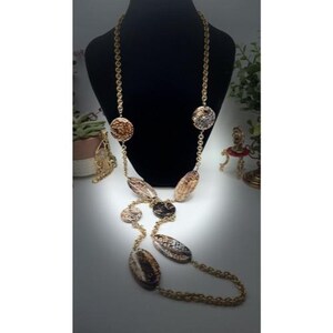 May include: A long gold-tone necklace with a variety of decorative elements. The necklace features a chain design with alternating round and oval-shaped pendants in shades of brown, beige, and black. The necklace is displayed on a black mannequin.