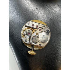 May include: Close-up of a vintage Omega watch movement. The gold-toned mechanism features intricate gears, ruby jewels, and the text "OMEGA WATCH Co. SWISS". The watch movement is round with a gold-colored outer ring and a winding stem.