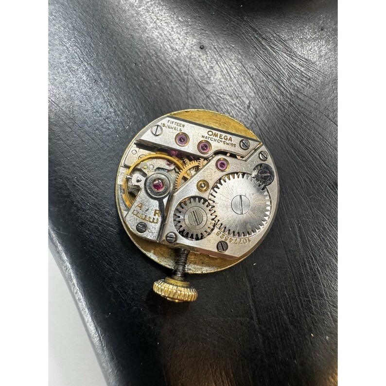 May include: Close-up of a vintage Omega watch movement. The gold-toned mechanism features intricate gears, ruby jewels, and the text "OMEGA WATCH Co. SWISS". The watch movement is round with a gold-colored outer ring and a winding stem.
