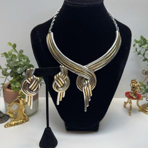 May include: A two-tone silver and gold-tone necklace and earring set. The necklace has a curved design with interwoven metal strands. The matching earrings are knot-shaped with dangling accents. The jewelry is displayed on a black mannequin.