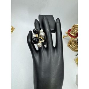 May include: A gold-toned ring featuring a cluster of spherical beads in varying sizes and colors, including glossy black, metallic silver, and small white pearls. The band is thin and embellished with small clear stones along the outer surface.