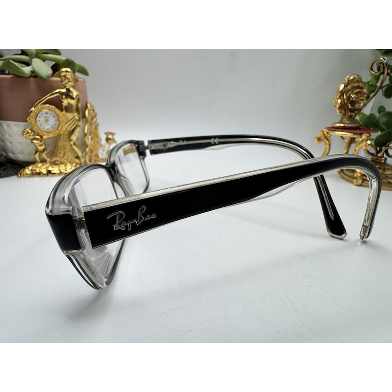 Puede incluir: Gafas con montura transparente y patillas negras. El logotipo de Ray-Ban es visible en la patilla izquierda. Las gafas tienen forma rectangular y est&aacute;n dise&ntilde;adas para la correcci&oacute;n de la visi&oacute;n. Las monturas son de material transparente y negro.