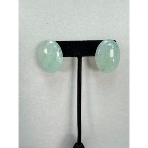 May include: A pair of oval-shaped, light green stone earrings with a smooth, polished finish. The earrings are mounted on a black T-shaped display stand, showcasing the translucent quality and subtle speckled texture of the pale green mineral.