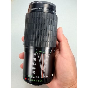 Könnte beinhalten: Ein schwarzes Takumar-A Zoom-Kameraobjektiv mit einem Brennweitenbereich von 70-200 mm und einer Blende von f/4. Der Tubus verfügt über einen strukturierten Gummigriff, weiße Entfernungsmarkierungen in Metern und Fuß sowie grüne Blendenwerte von f/4 bis f/32.