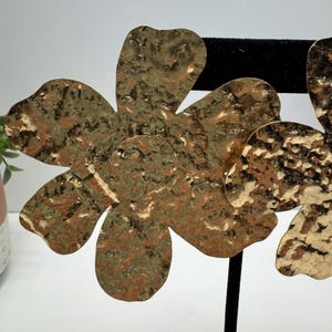 May include: Large, hammered-metal flower earrings in shades of gold and bronze. The earrings are displayed on a black stand. The flower design has five petals with a textured surface. The earrings are a statement piece.