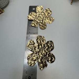 May include: Two gold-colored, hammered-metal flower-shaped embellishments. A ruler shows the flowers are approximately 3 inches in diameter. The flowers have a textured surface and five petals. The background is a light gray.