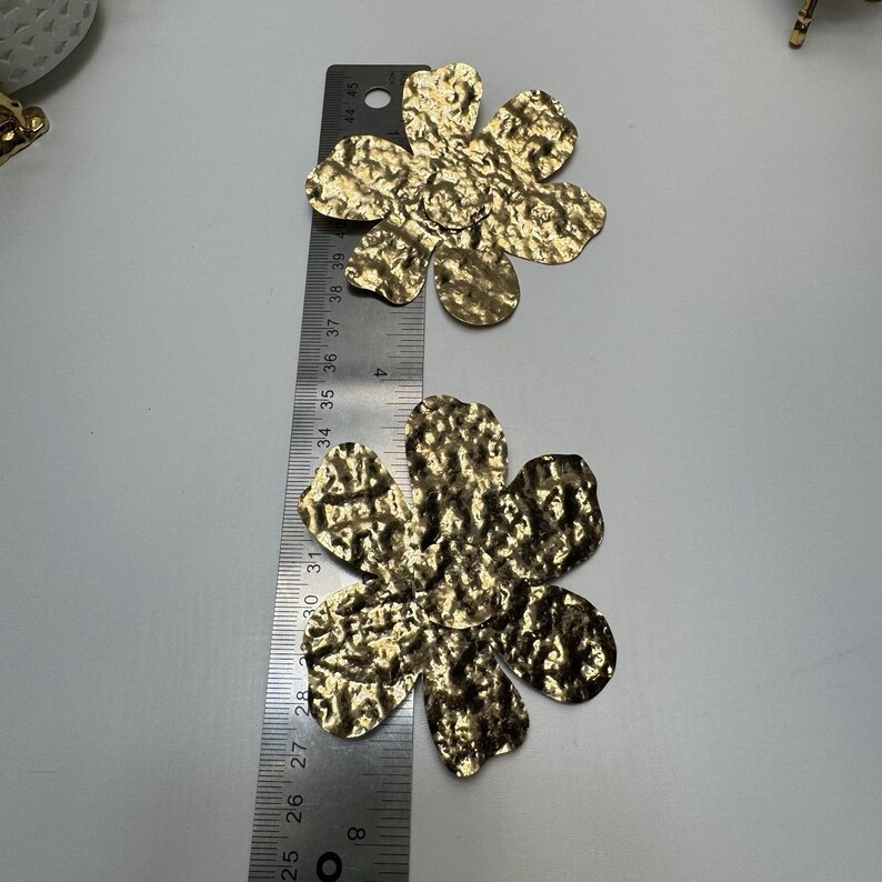 May include: Two gold-colored, hammered-metal flower-shaped embellishments. A ruler shows the flowers are approximately 3 inches in diameter. The flowers have a textured surface and five petals. The background is a light gray.