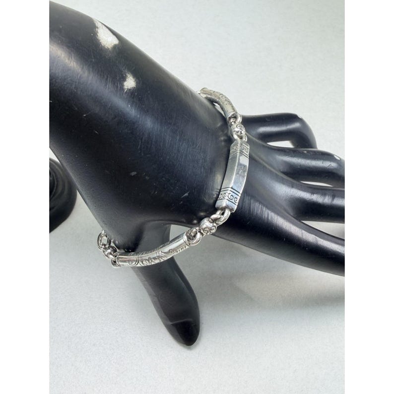 K&ouml;nnte beinhalten: Ein silbernes Armband mit aufwendigen Gravuren und einem rechteckigen Mittelst&uuml;ck. Das Armband wird auf einer schwarzen Mannequin-Hand pr&auml;sentiert, um seine detaillierte Verarbeitung und seinen eleganten Stil hervorzuheben. Die Glieder des Armbands sind mit Mustern verziert.
