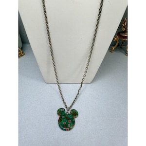 May include: A silver-toned chain necklace with a green pendant in the shape of a mouse head. The pendant is decorated with small red and white floral patterns. The necklace is displayed on a white mannequin.