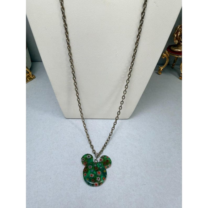May include: A silver-toned chain necklace with a green pendant in the shape of a mouse head. The pendant is decorated with small red and white floral patterns. The necklace is displayed on a white mannequin.