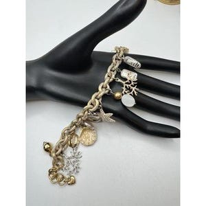 May include: A gold-toned charm bracelet featuring various ocean-themed charms. Charms include a starfish, seashell, coral, and a snowflake. The bracelet is displayed on a black hand against a white background.