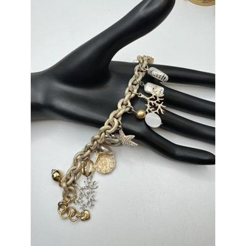 May include: A gold-toned charm bracelet featuring various ocean-themed charms. Charms include a starfish, seashell, coral, and a snowflake. The bracelet is displayed on a black hand against a white background.