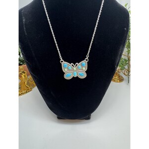 May include: A silver-toned butterfly pendant necklace. The butterfly pendant features turquoise and light green stones. The necklace is displayed on a black velvet bust. The background is white.