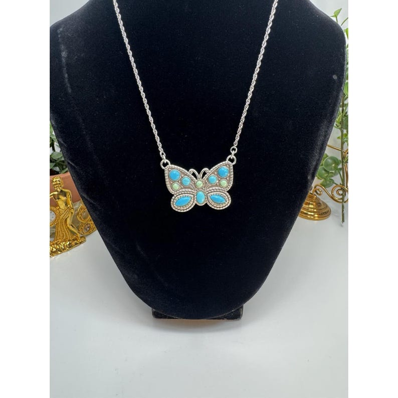 May include: A silver-toned butterfly pendant necklace. The butterfly pendant features turquoise and light green stones. The necklace is displayed on a black velvet bust. The background is white.
