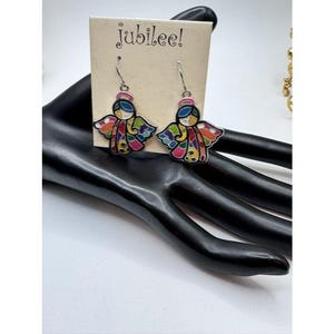 May include: A pair of dangle earrings featuring colorful angel designs. Each earring has a silver hook and a detailed angel figure with a pink halo, blue hair, and multi-colored wings. The earrings are displayed on a black hand-shaped jewelry holder, with a tag that reads "jubilee!"