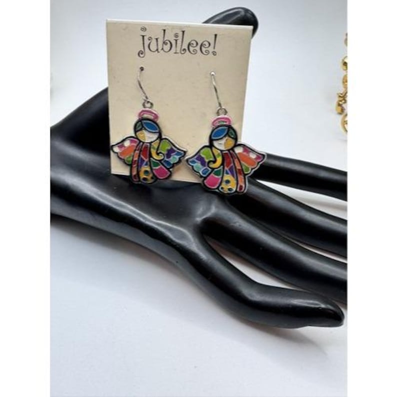 May include: A pair of dangle earrings featuring colorful angel designs. Each earring has a silver hook and a detailed angel figure with a pink halo, blue hair, and multi-colored wings. The earrings are displayed on a black hand-shaped jewelry holder, with a tag that reads "jubilee!"