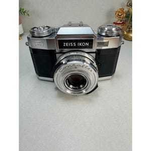 May include: A silver and black Zeiss Ikon Contaflex camera featuring a prominent circular lens with the text Contaflex and Zeiss Tessar printed on the front. The camera body includes metallic dials on the top and a black textured grip.