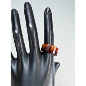 May include: A wide band ring featuring a glossy tortoiseshell pattern with blended shades of dark brown, amber, and black. The ring has a smooth, polished finish and a uniform thickness throughout the entire circumference of the circular band.