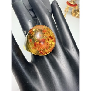 May include: A large, round amber ring with red and yellow inclusions. The ring has a clear band and is set on a black surface. The amber is translucent, allowing light to pass through.