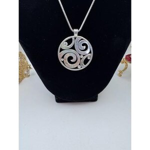 以下が含まれることがあります： 渦巻き模様と小さな透明な宝石をあしらった、円形デザインのシルバーペンダントネックレス。ペンダントは繊細なシルバーチェーンから吊り下げられています。ネックレスは黒いベルベットの表面に展示されています。
