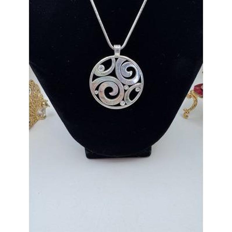 以下が含まれることがあります： 渦巻き模様と小さな透明な宝石をあしらった、円形デザインのシルバーペンダントネックレス。ペンダントは繊細なシルバーチェーンから吊り下げられています。ネックレスは黒いベルベットの表面に展示されています。