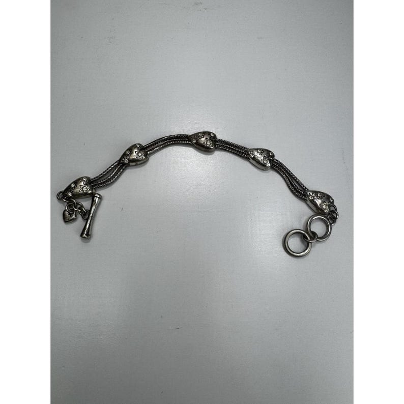 May include: Silver bracelet with heart-shaped charms on a multi-strand chain. The bracelet has a toggle clasp and two decorative rings. The hearts are decorated with small, sparkling details. A charming piece of jewellery.