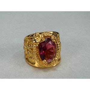 May include: A gold-toned ring featuring a large, oval-cut, deep red gemstone set in the center. The band is crafted with a textured, ornate metal surface that surrounds the stone, showcasing intricate detailing and a polished finish throughout.