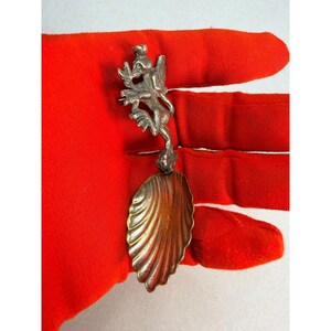 May include: A silver-toned metal spoon featuring a decorative handle shaped like a winged mythical creature. The bowl of the spoon has a distinct shell-like design with vertical ridges radiating from the base of the handle to the rounded edge.