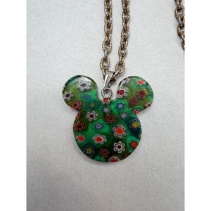 May include: A green Mickey Mouse-shaped pendant necklace with a floral design. The pendant is made of glass and features a pattern of small, colorful flowers. The chain is a silver-colored metal. The necklace is a fun and whimsical accessory.
