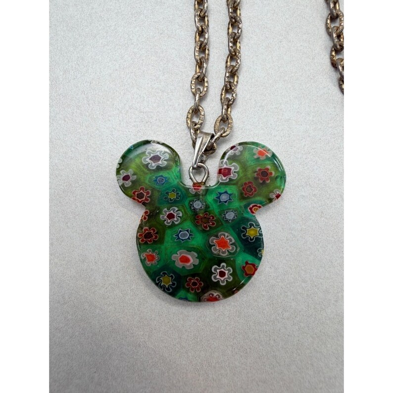 May include: A green Mickey Mouse-shaped pendant necklace with a floral design. The pendant is made of glass and features a pattern of small, colorful flowers. The chain is a silver-colored metal. The necklace is a fun and whimsical accessory.