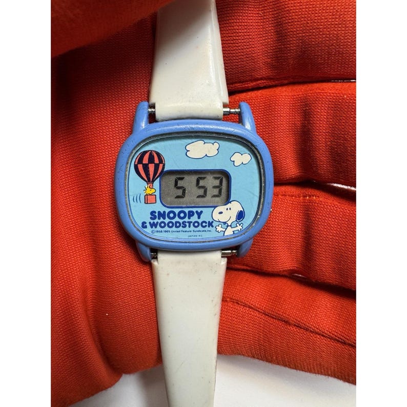 May include: A vintage Snoopy & Woodstock digital watch with a blue face and white band. The watch face features Snoopy, Woodstock, a hot air balloon, and the time 5:53. The watch band is white.