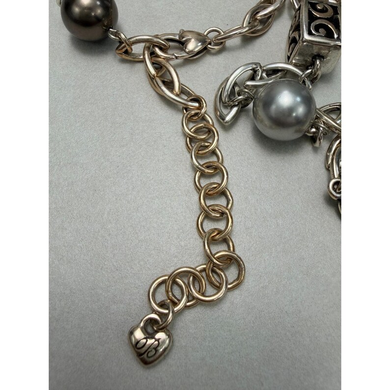 Może przedstawiać: Srebrny naszyjnik z mieszanką styl&oacute;w łańcuszk&oacute;w i element&oacute;w dekoracyjnych. Naszyjnik posiada zawieszkę w kształcie serca z inicjałami "B" i "G", a także r&oacute;żnorodne koraliki i ogniwa. Naszyjnik posiada zapięcie na karabińczyk.