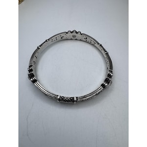 May include: Silver-toned bangle bracelet with intricate detailing. The bracelet features a series of decorative elements, including floral and geometric patterns. The design incorporates multiple bands and textured surfaces, creating a detailed and ornate appearance.