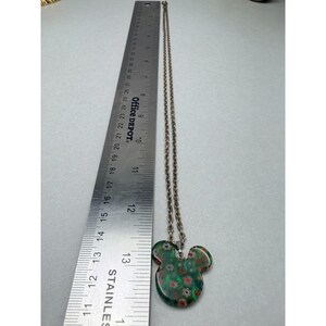 May include: A silver-toned necklace with a pendant shaped like Mickey Mouse's head. The pendant is green with red, white, and blue floral patterns. The ruler next to the necklace shows the pendant is approximately 1.5 inches wide.