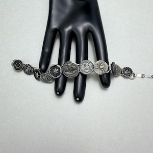 May include: A silver bracelet featuring a series of vintage buttons with various designs, including a ship, a star, and floral patterns. The bracelet is displayed on a black hand-shaped form against a white background.
