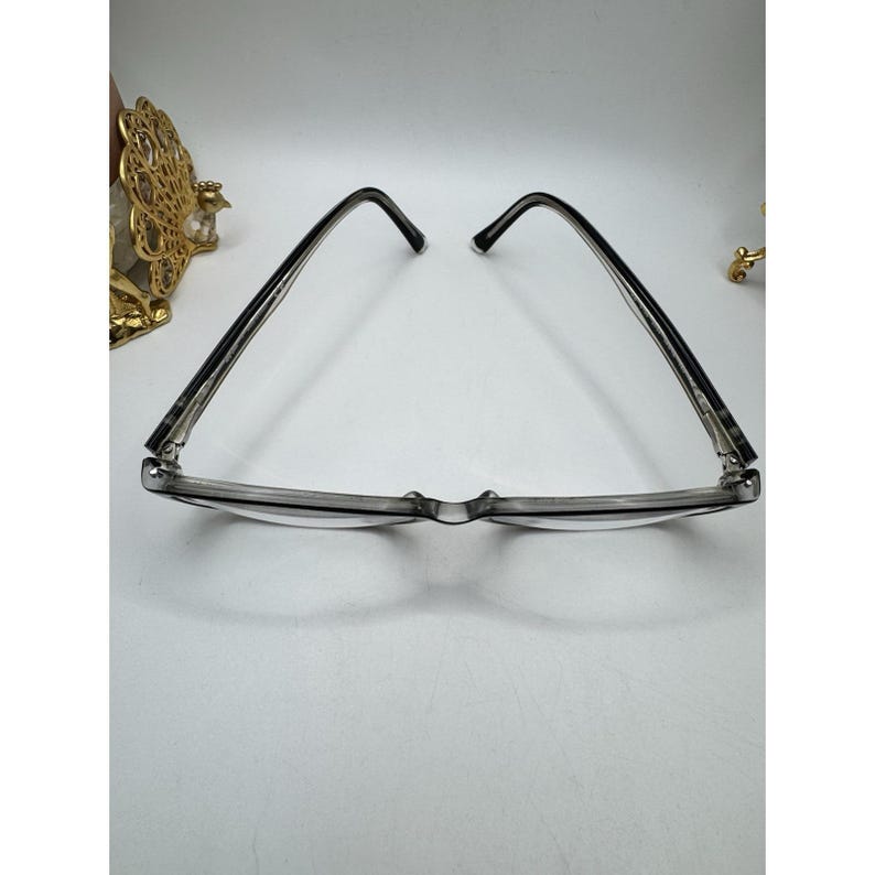 Puede incluir: Un par de gafas con montura transparente, varillas y puente de color gris oscuro. Las gafas est&aacute;n sobre un fondo blanco, con un objeto decorativo dorado visible en el lado izquierdo.