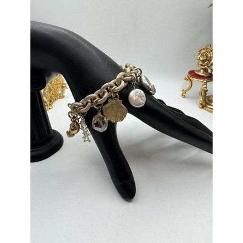 May include: A gold-toned charm bracelet with various charms, including a seashell, a pearl, and other decorative elements. The bracelet is displayed on a black hand-shaped display stand against a white background. The bracelet's chain is thick and textured.